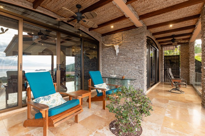 Patio with blue cushioned chairs, stone wall, and ocean view through glass doors.
