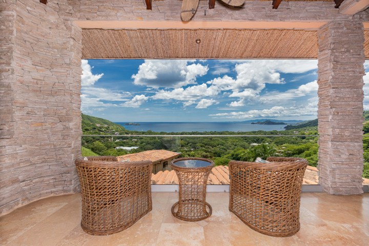 Patio with wicker chairs and table overlooking a lush landscape and ocean view under a cloudy blue sky.