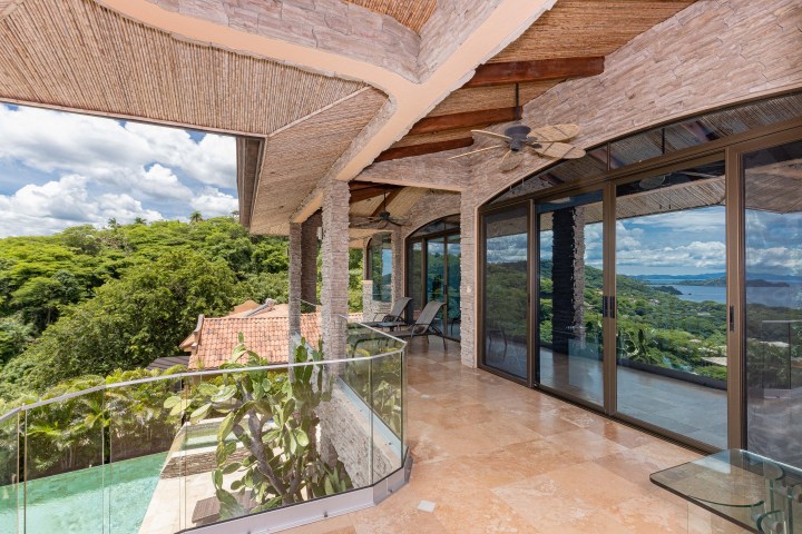 Outdoor patio with glass railing and ocean view, featuring stone walls and ceiling fan.