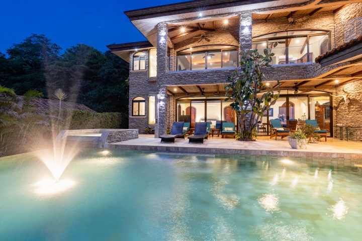 Luxurious house with pool and fountain illuminated at night, surrounded by trees.