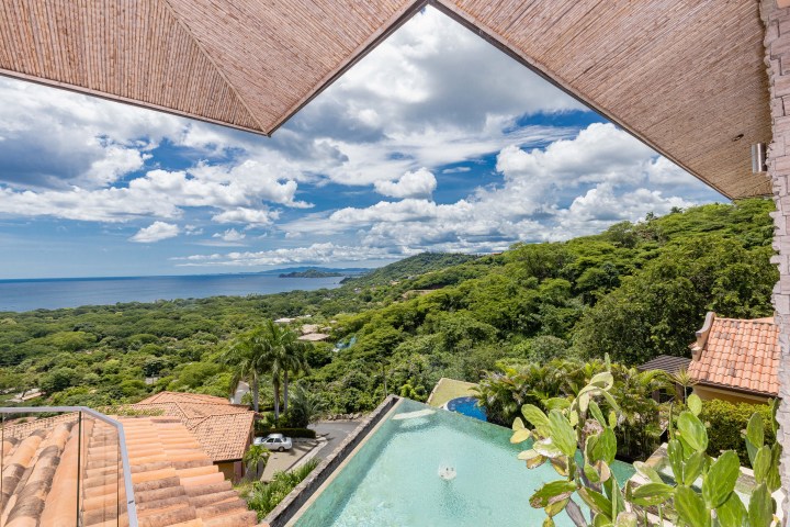 Coastal view with infinity pool, lush greenery, and ocean under a partly cloudy sky.