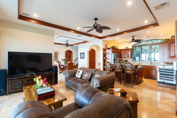 Spacious open-plan living room with brown sofas, TV, kitchen area with bar stools, and decorative ceiling fans.