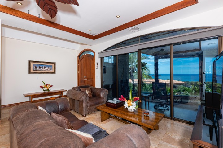 Living room with brown sofas, wooden table, large window showing ocean view, and decorative ceiling fan.