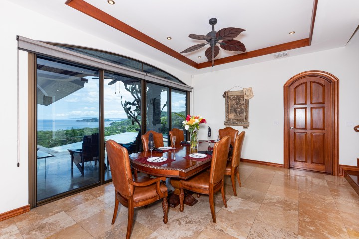 Elegant dining room with ocean view, wooden table, and leather chairs under a fan with leaf-shaped blades.