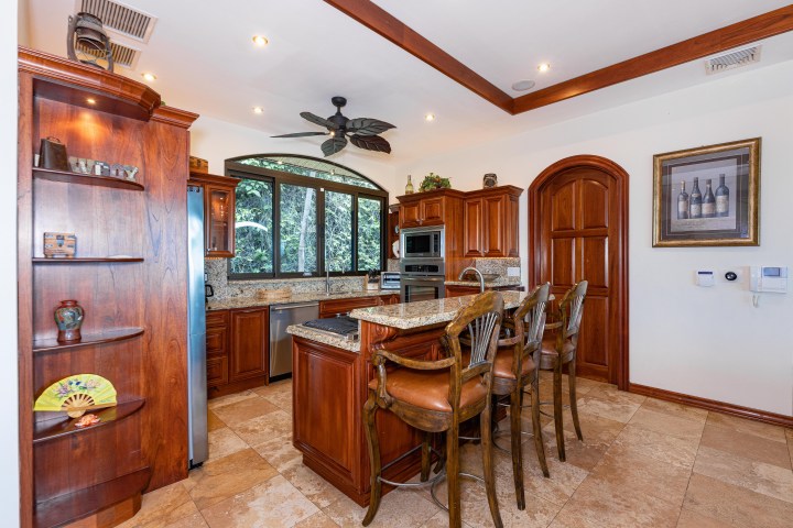 Warm kitchen with wood cabinets, marble countertops, bar stools, and large window overlooking greenery.