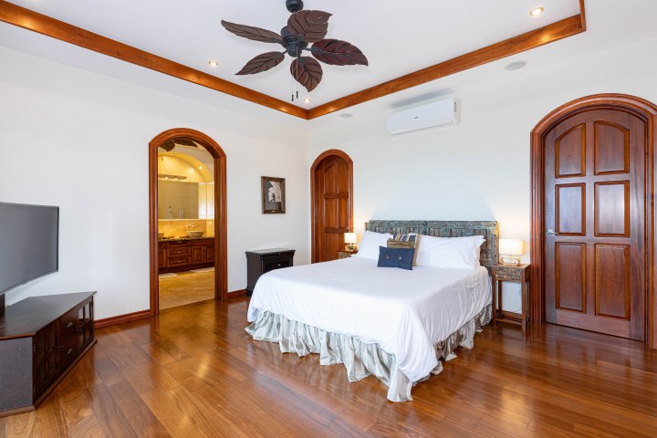 Spacious bedroom with wooden floor, bed, and en-suite bathroom, featuring ceiling fan and wooden doors.