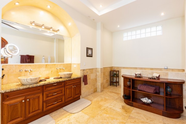 Spacious bathroom with double sinks, wooden cabinets, large mirror, and shelf unit on tile floor.