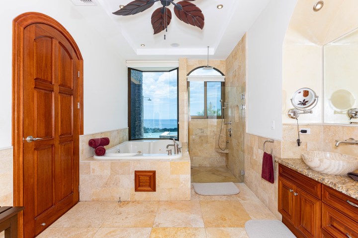 Luxurious bathroom with tub, large window view, shower, wooden door, and beige tiles.