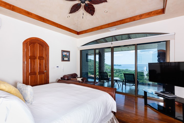 Bedroom with wooden ceiling and door, ocean view through large glass doors, TV on the right.