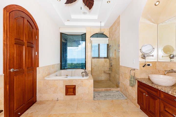 Luxurious bathroom with wooden door, corner bathtub, large shower, and ocean view through glass window.
