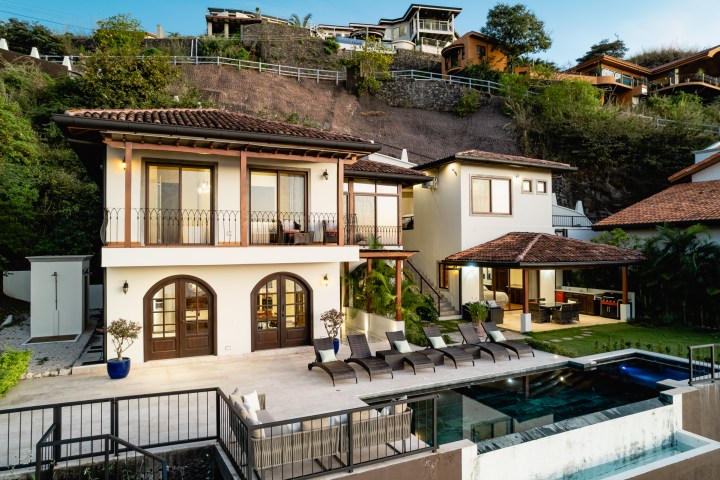 Modern hillside villa with pool, patio, and lounge chairs, surrounded by lush greenery and neighboring homes.