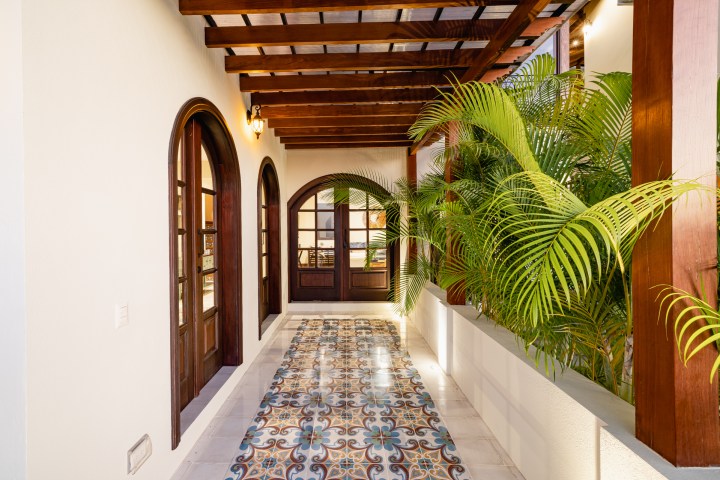 Hallway with arched wooden doors, patterned tiles, and potted palm plants under wooden beams.
