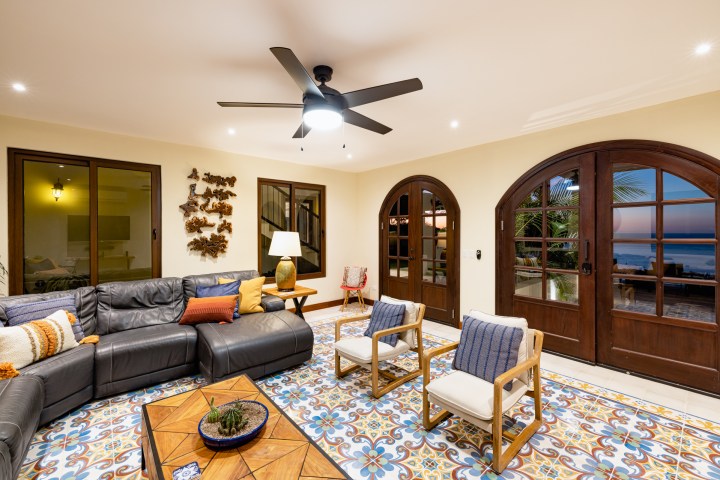 Living room with tiled floor, dark sofa, two chairs, ceiling fan, and ocean view through arched windows.