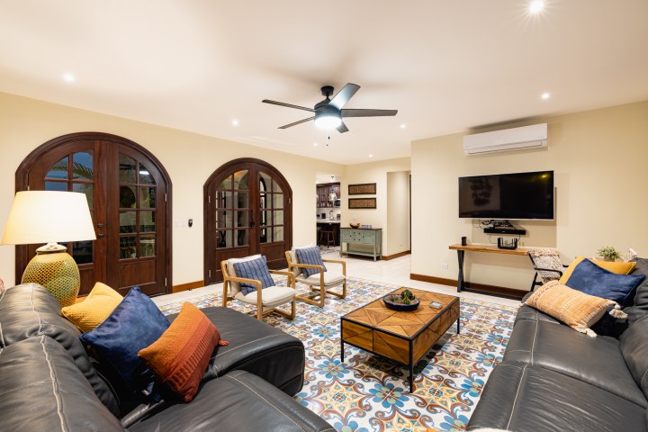 Spacious living room with leather sofas, colorful pillows, wooden doors, ceiling fan, and wall-mounted TV.