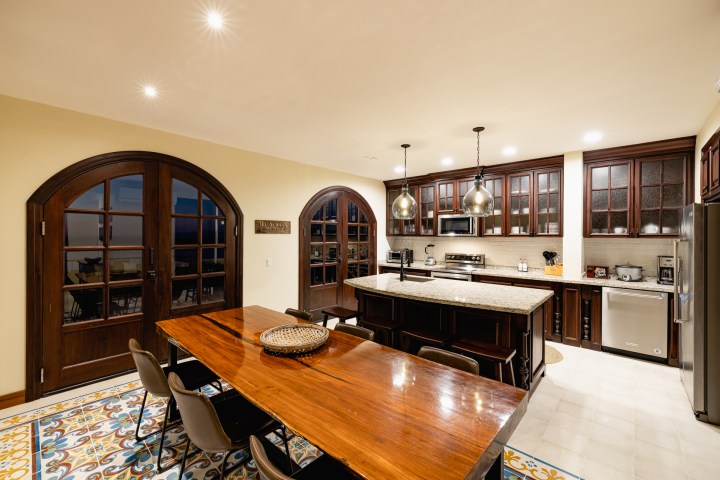 Spacious kitchen with a long wooden table, tiled floor, and dark wooden cabinets, illuminated by pendant lights.