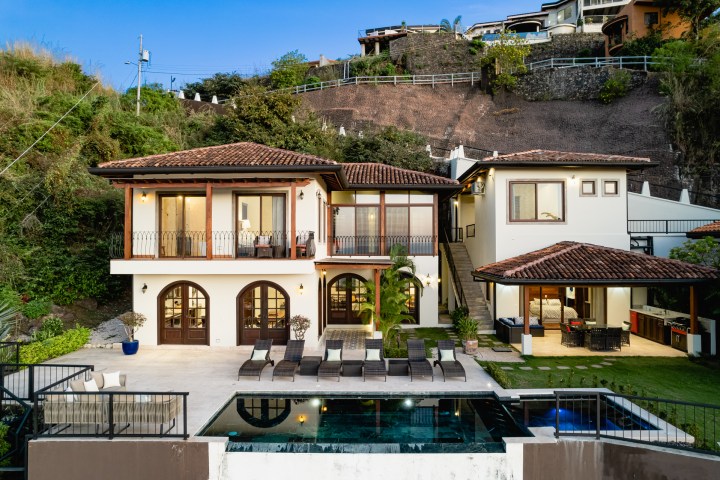 Two-story house with pool, patio, and hillside view at dusk.