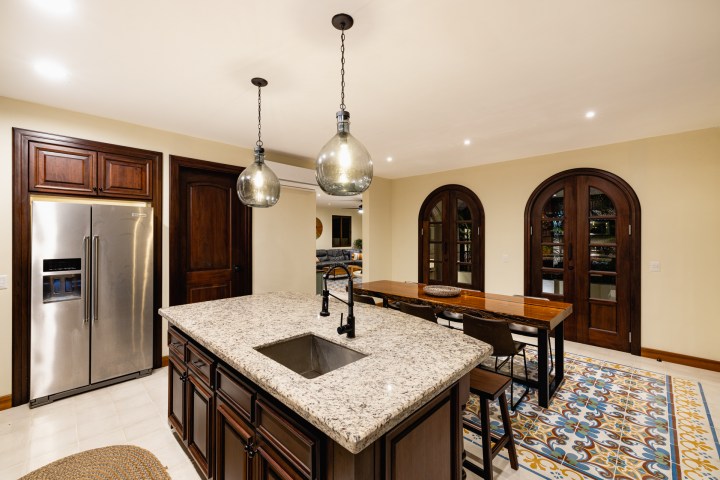 Modern kitchen with granite island, hanging lights, stainless fridge, and arched wooden doors.