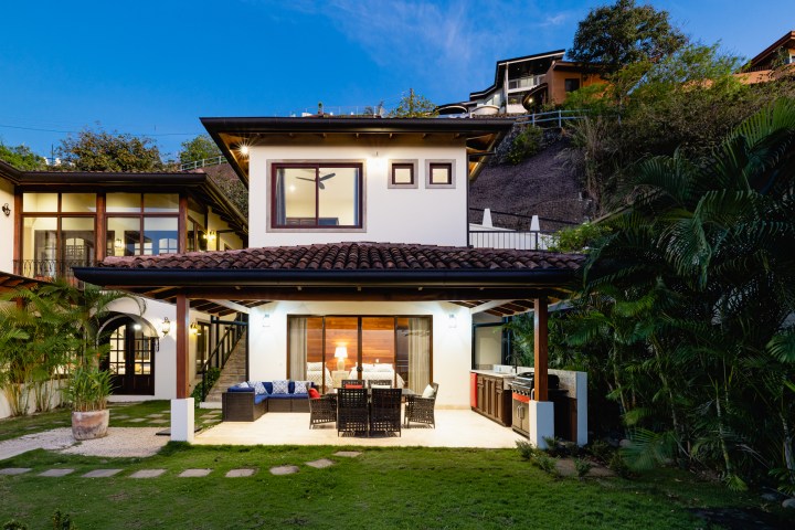 Two-story house with outdoor patio and furniture, set in a lush garden environment, during early evening.