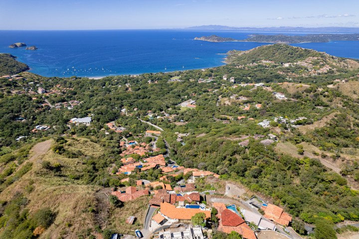 Aerial view of coastal landscape with red-roofed buildings, green hills, and blue ocean.