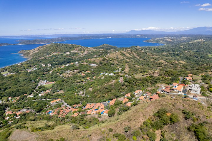 Aerial view of a coastal town with lush hills and blue ocean under a clear sky.
