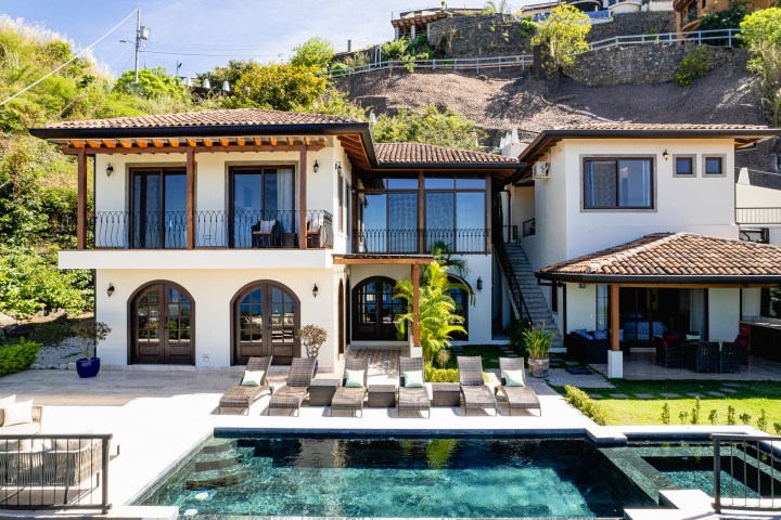 Two-story villa with pool, loungers, and greenery on hillside background.