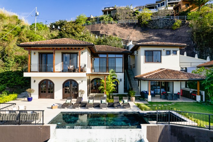 Luxury villa with two floors, patio, pool, and hillside in the background under a clear blue sky.