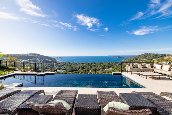 Infinity pool with ocean view, surrounded by lounge chairs and sofa under blue sky.