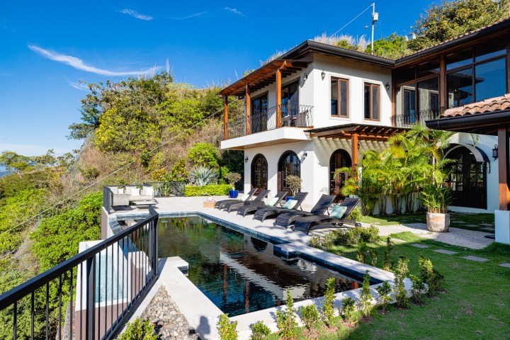 Modern house with a pool, sun loungers, and greenery on a hillside under a clear blue sky.