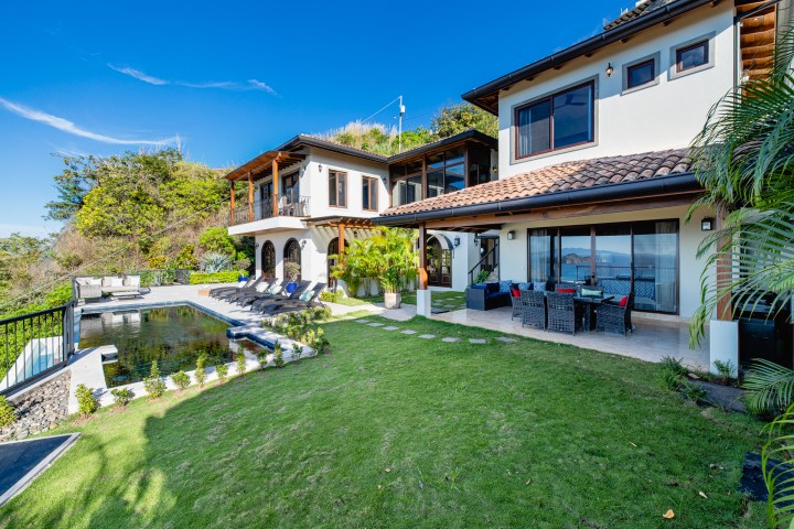 Modern hillside house with pool, patio, and lush garden under a clear blue sky.