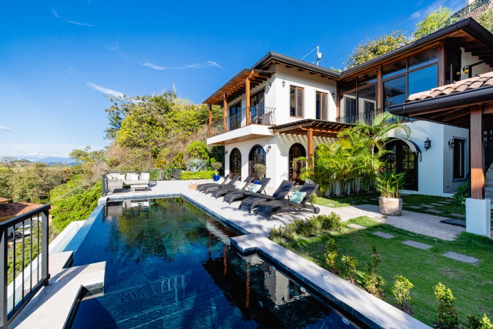 Luxury villa with pool, sun loungers, and lush greenery under a clear blue sky.