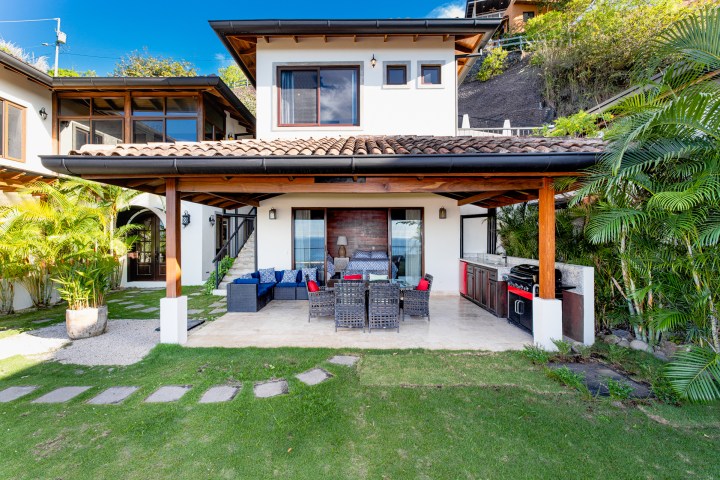 Modern house with patio, outdoor seating, grill, and lush greenery in yard.