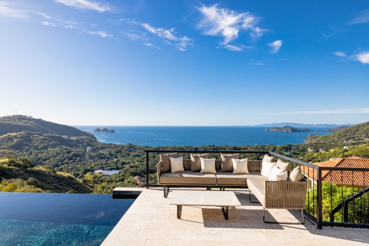 Outdoor seating area with a couch overlooking a scenic ocean view and lush green hills.