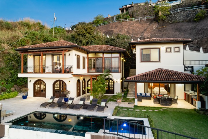 Modern two-story villa with pool, patio, and lawn on a hillside.