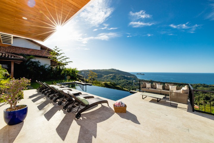 Sunlit patio with loungers, pool, and ocean view under a clear blue sky.