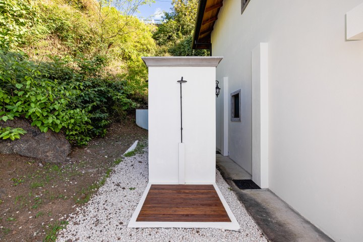 Outdoor shower with white walls and wooden floor, beside a white building and greenery.