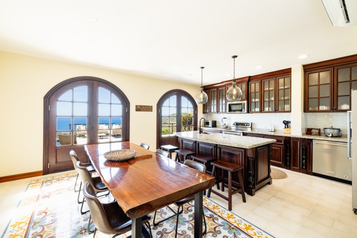 Bright kitchen with wood cabinets, large dining table, and ocean view through arched windows.