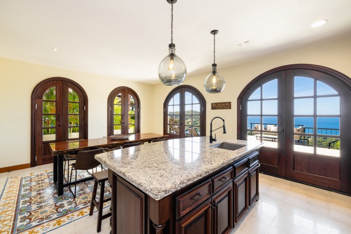 Bright kitchen with granite island, pendant lights, arched doors, ocean view, and dining table on patterned floor.