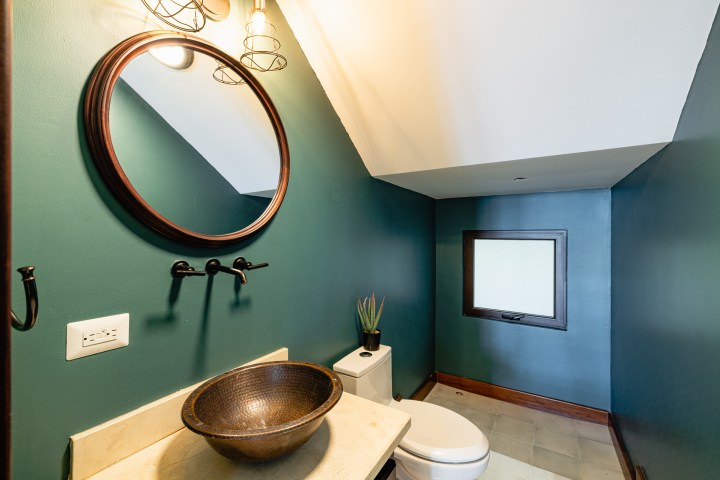 Compact bathroom with a round mirror, vessel sink, and potted plant on a teal wall.