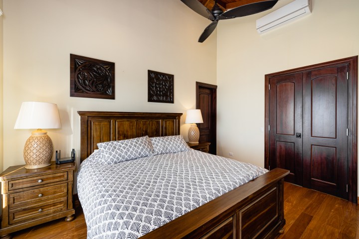Bedroom with a wooden bed, patterned bedding, nightstands, lamps, ceiling fan, and decorative wall panels.