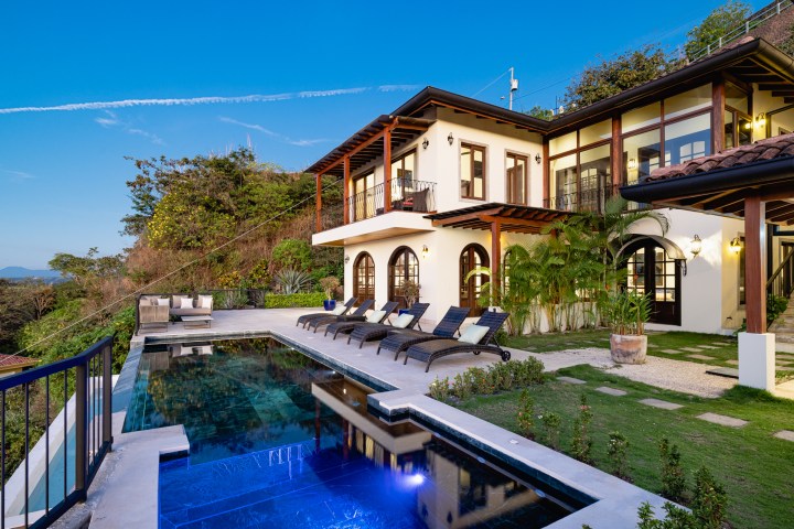 Luxurious villa with pool, loungers, and lush greenery against a clear blue sky.