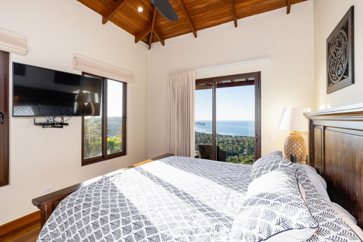 Bedroom with sea view, wooden ceiling, TV, patterned bedding, and large windows with curtains.