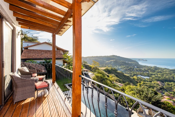 Balcony view with wicker chairs overlooking hills, ocean, and houses under a bright sky.