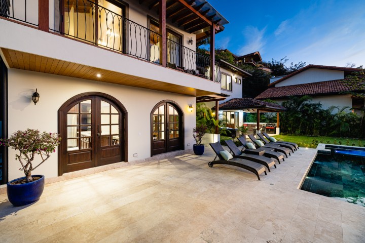 Luxury villa patio at dusk with loungers, pool, and greenery.
