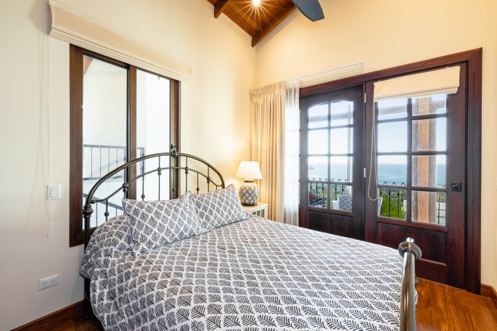 Bright bedroom with a patterned bedspread, large windows, and a view of the ocean.