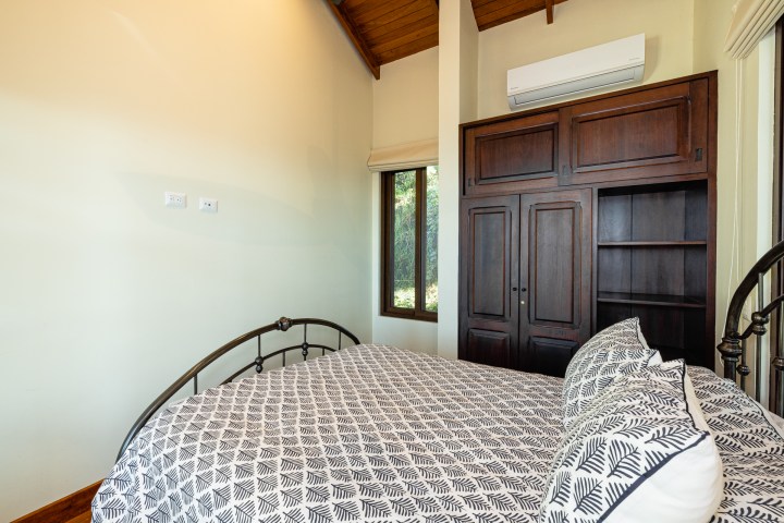 Bedroom with patterned bedspread, wood wardrobe, air conditioner, and a window with greenery outside.