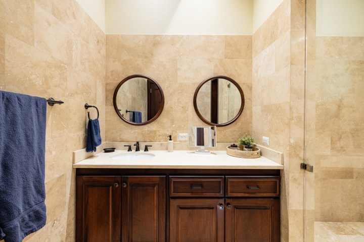 Bathroom with beige tiles, dark wood vanity, round mirrors, and blue towels hanging.