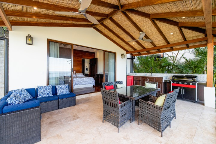 Covered patio with wicker furniture, glass dining table, outdoor kitchen, and adjacent bedroom with sliding doors.