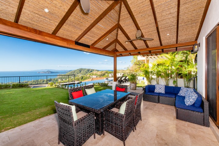 Patio with blue cushions, wicker furniture, ocean view, wooden roof, and green lawn.