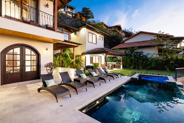 Luxurious villa with a pool, patio, lounge chairs, and lush greenery under a clear blue sky.