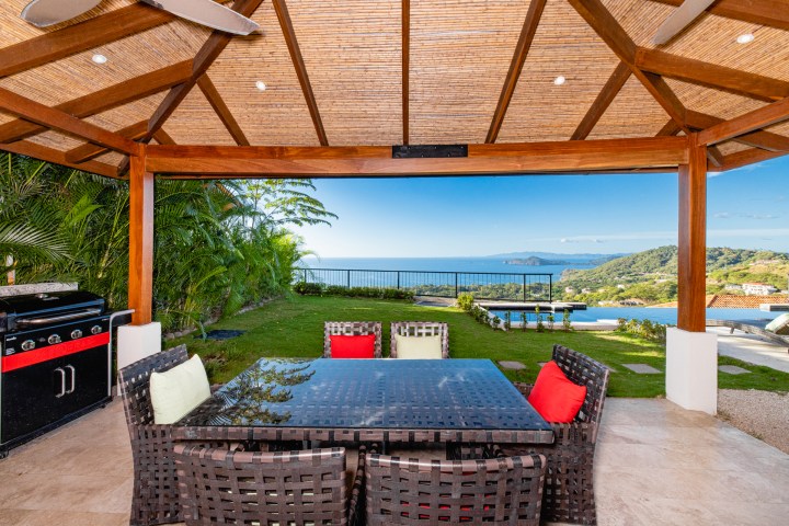 Outdoor patio with wicker furniture, grill, and ocean view under a wooden pergola.